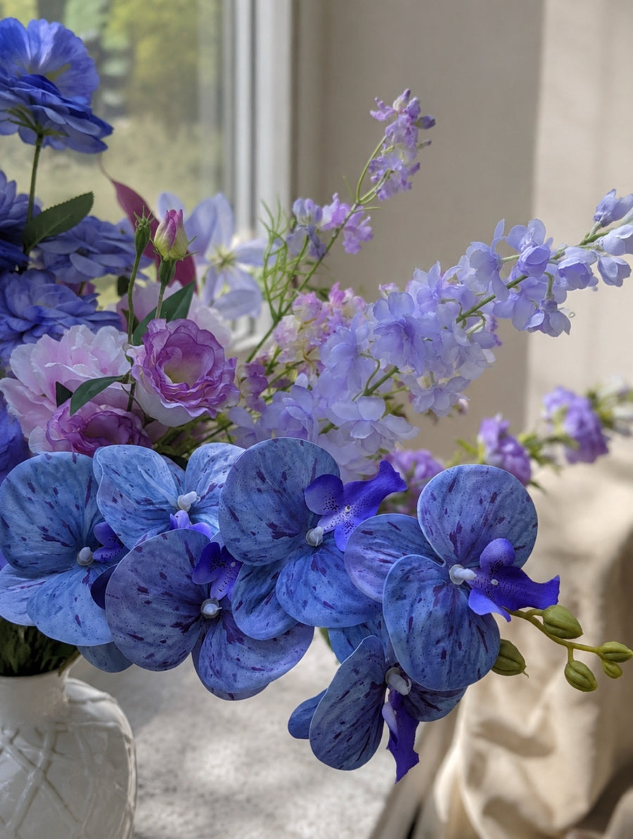 Close-up of the Water Lily Collection artificial floral arrangement showcasing pastel blue hydrangea-inspired blooms, lavender florals and blush pink accents in a white ceramic vase.